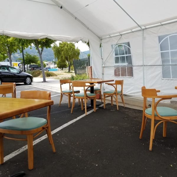Outdoor dining area under a white tent with wooden tables and chairs, cars parked nearby.