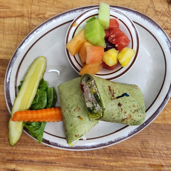 A Mother Hubbard's breakfast plate with a green wrap, fruit cup, pickle spear, and carrot.