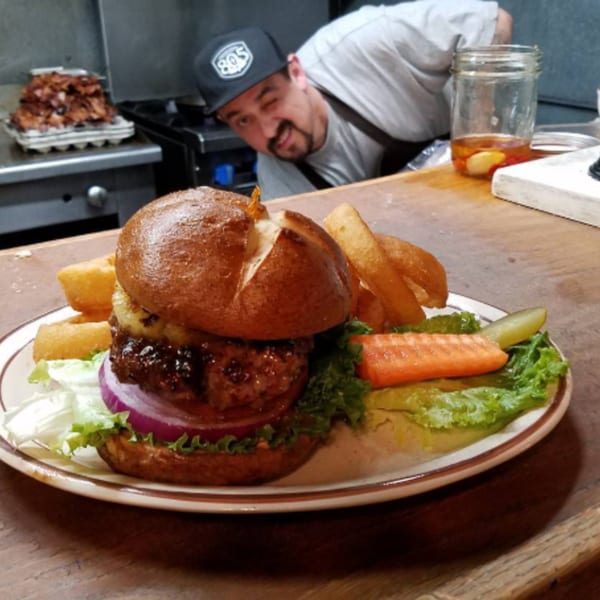 A chef winks from the kitchen behind a burger with fries, lettuce, onion, pickle, and carrot.